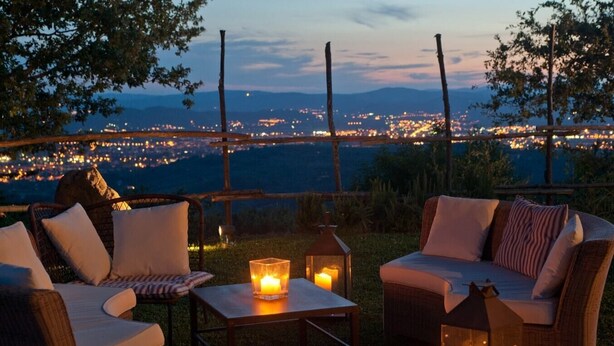 Uma área de estar ao ar livre com móveis de vime e velas, com vista para um horizonte urbano ao entardecer.