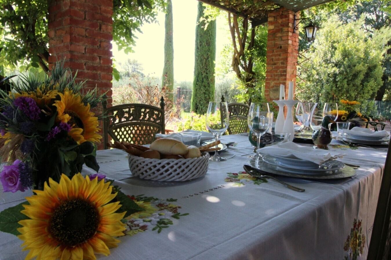 Un área de comedor al aire libre con una mesa preparada para dos personas, rodeada de follaje y un muro de ladrillo.