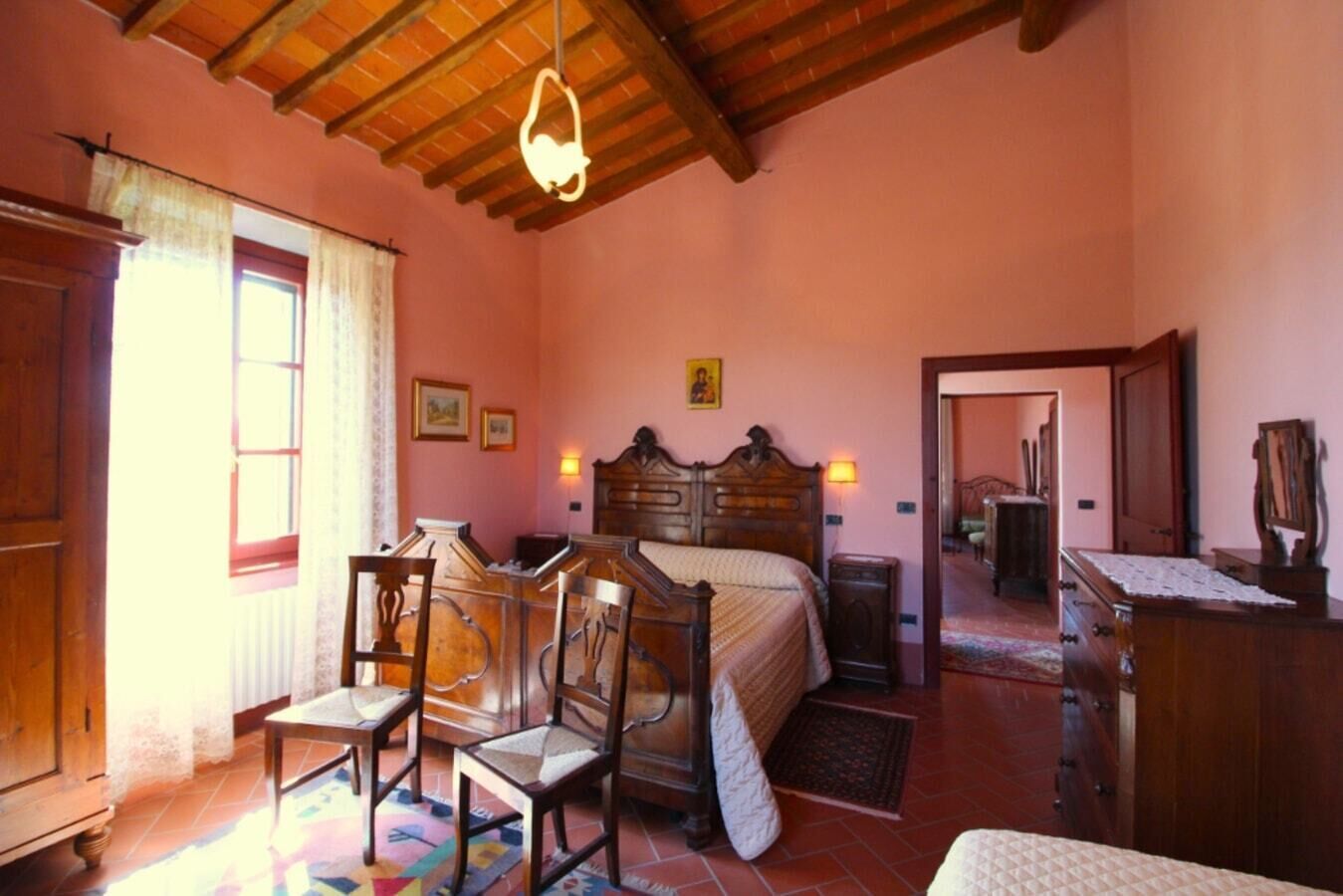 A bedroom with a wooden bed, a desk, and chairs, a window with curtains, and a door leading to another room.