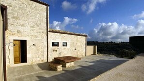 Un edificio de piedra con puerta y ventanas de madera, un patio de concreto con una mesa de madera y vistas a un paisaje montañoso.