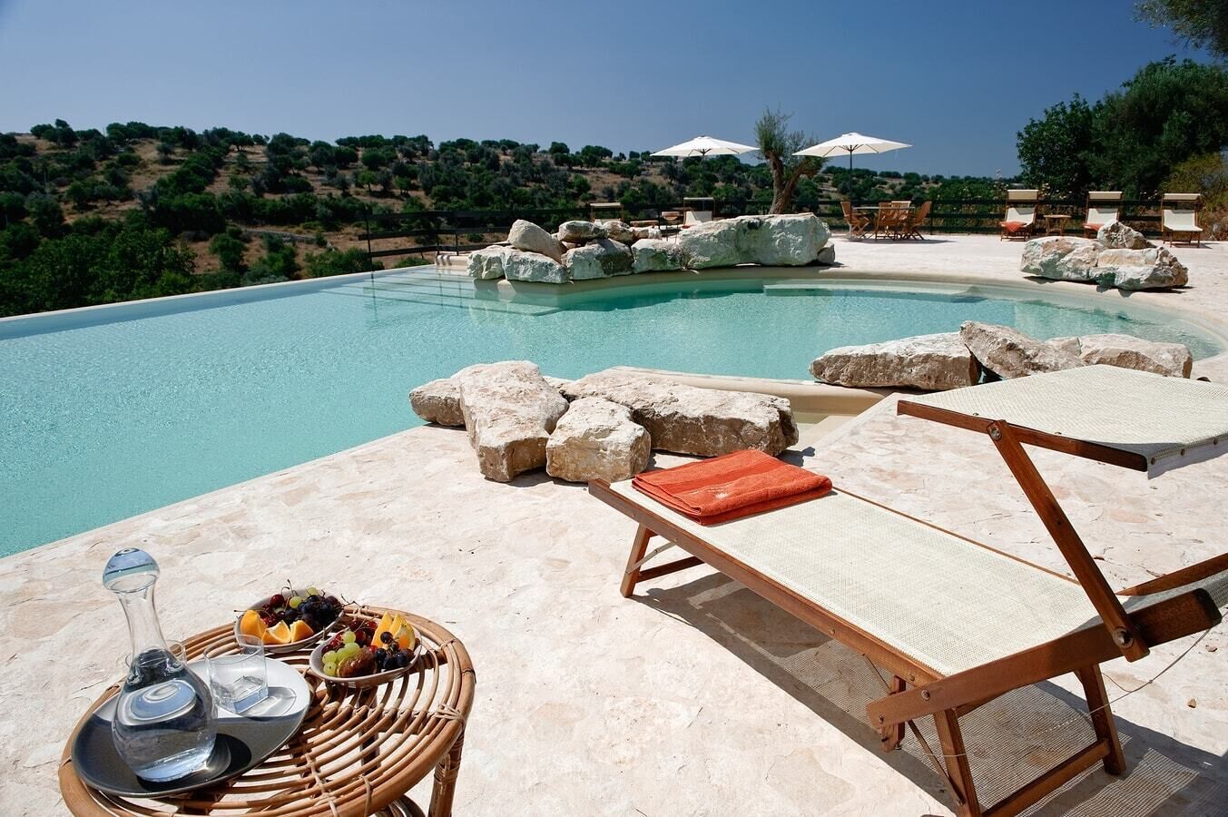 A poolside area with lounge chairs, a table with a fruit basket, and a view of the surrounding landscape.