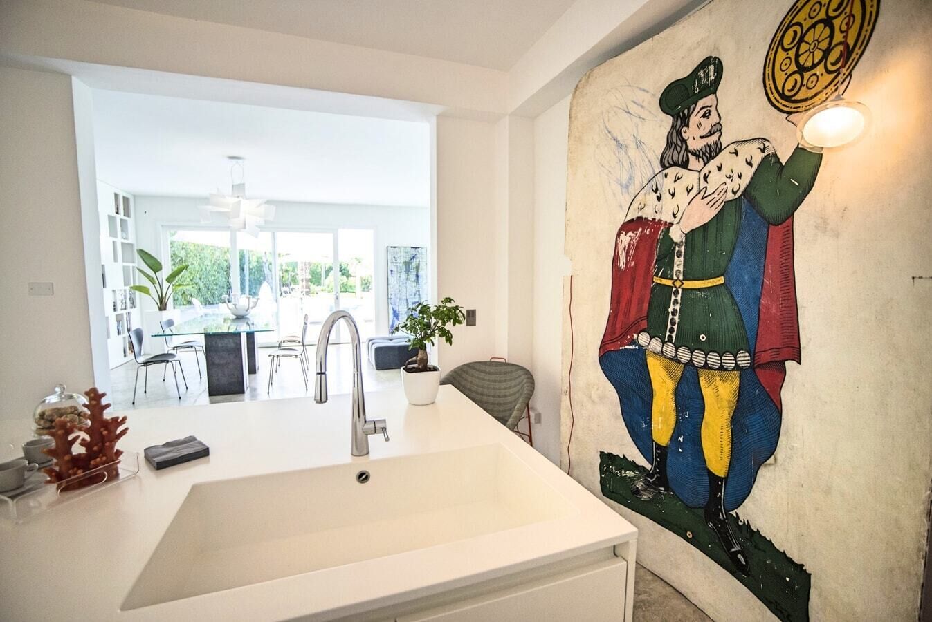 Una cucina moderna con un lavello bianco capiente, una zona pranzo con tavolo e sedie in vetro, e un murale sulla parete.