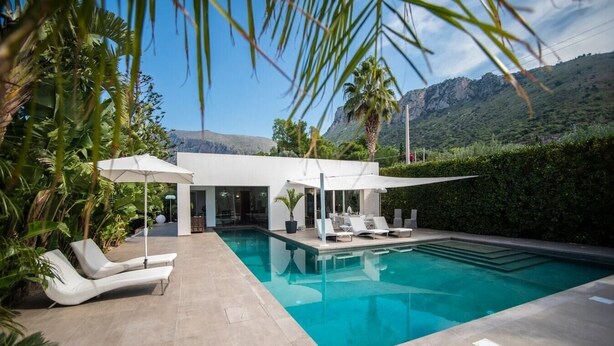 A modern pool area with lounge chairs and umbrellas, surrounded by lush greenery and a mountain backdrop.