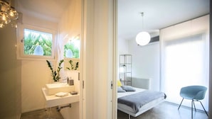A modern bedroom with a bed, bedside table, and a chair. There is a window with a view of greenery outside.