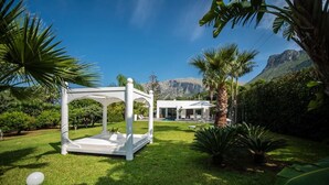 A white gazebo in a lush garden with a pool and a modern house in the background.