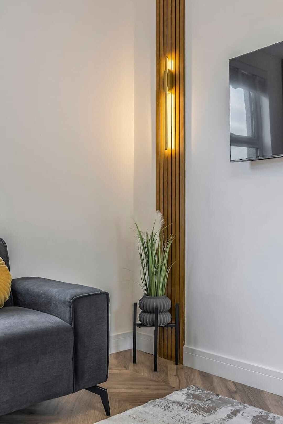 A modern living room with a grey sofa, a wooden accent wall, a small black chair, a potted plant, and a flat-screen TV mounted on the wall.