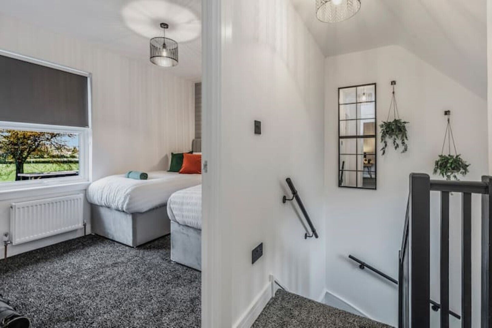 Une chambre d’hôtel avec une grande fenêtre, un radiateur, un lit avec des oreillers, une table de chevet et une plante suspendue.