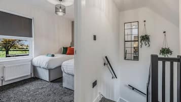 Une chambre d’hôtel avec une grande fenêtre, un radiateur, un lit avec des oreillers, une table de chevet et une plante suspendue.