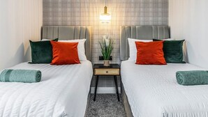 Hotelkamer met twee bedden, elk met witte beddengoed en kussens in groen en oranje. Daartussen staat een kleine tafel met een lamp en een plant.
