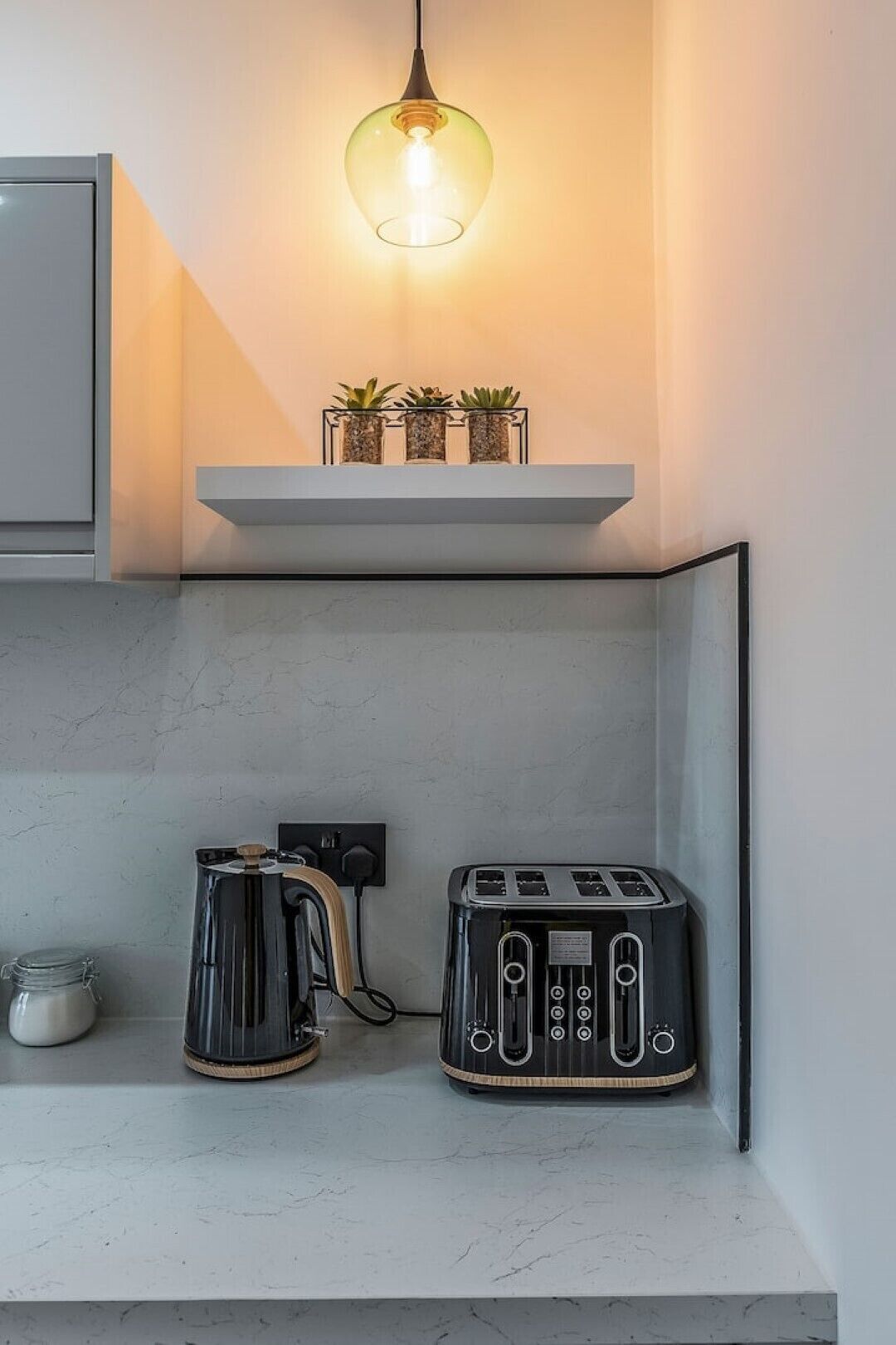 A modern kitchen with a coffee maker, toaster, and a hanging light fixture above a shelf with plants.