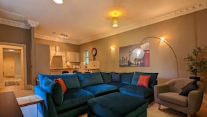A living room with a sectional sofa, a small table, a chair, and a kitchen in the background. - Stunning & Modern 2BD Flat - Manchester! (Salford)