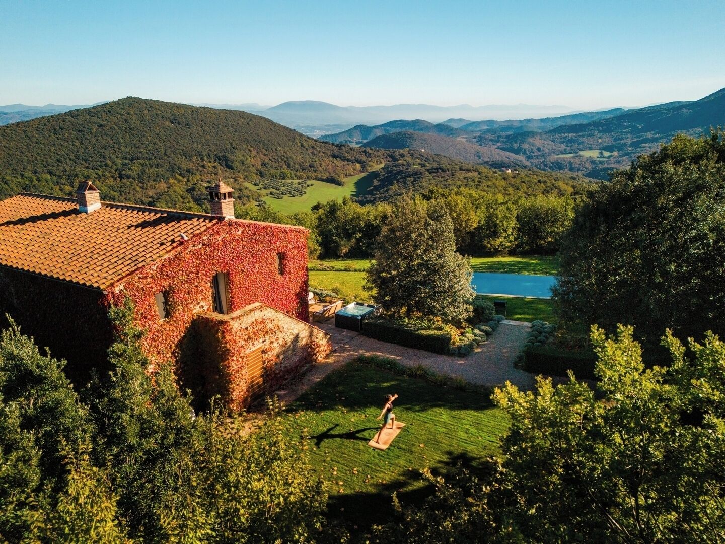 A rustic stone building with a tiled roof, surrounded by lush greenery and a clear view of a lake and distant hills.
