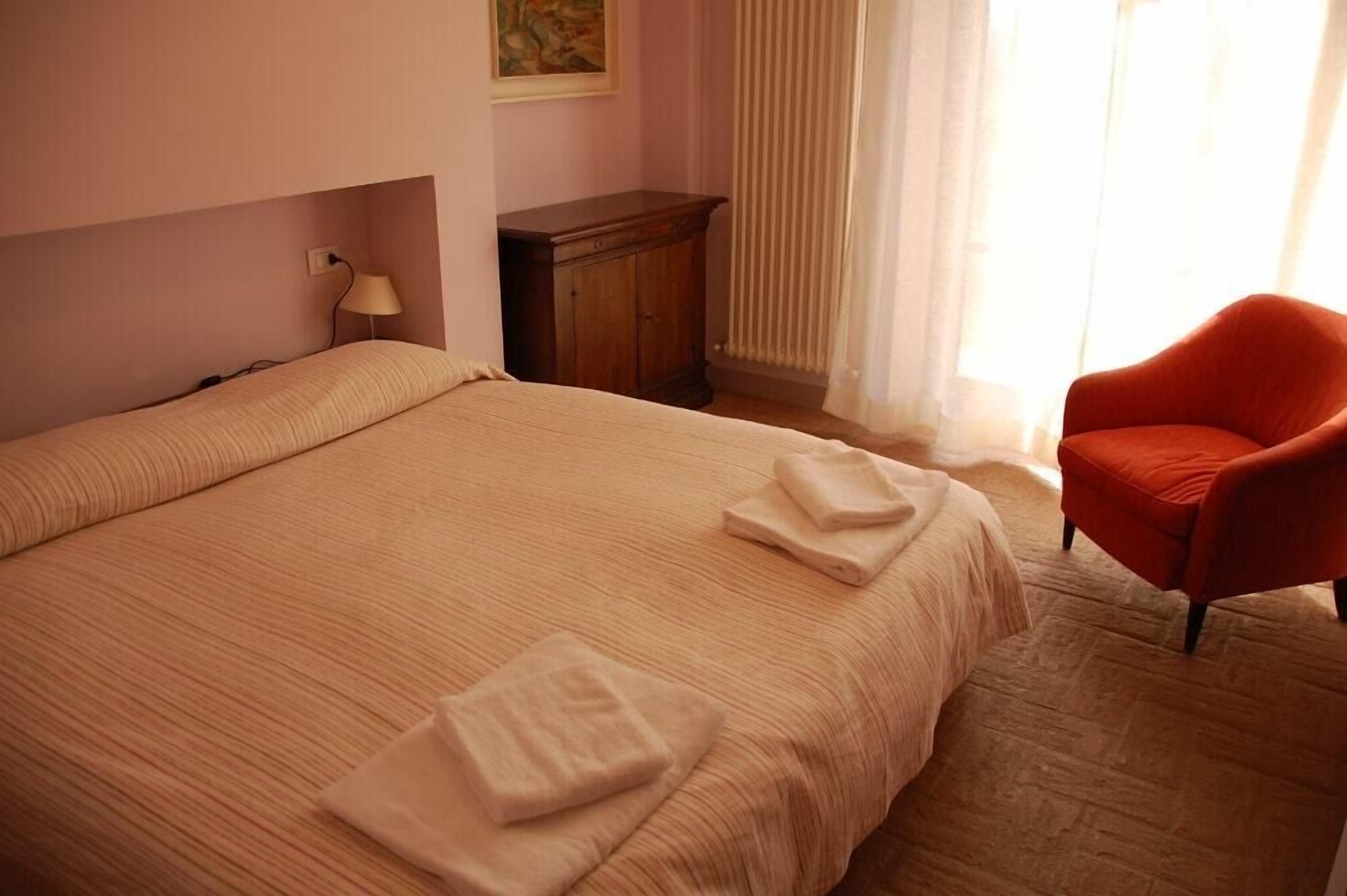 A neatly made bed with two folded towels, a wooden nightstand with a lamp, and a red armchair in a room with light pink walls and a window with sheer curtains.