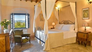 A four-poster bed with white curtains, a green armchair, a wooden nightstand with a lamp, and a view of the outdoors through a window. - Villa Bibbona - Private villa with heated pool (Montecatini Val di Cecina)