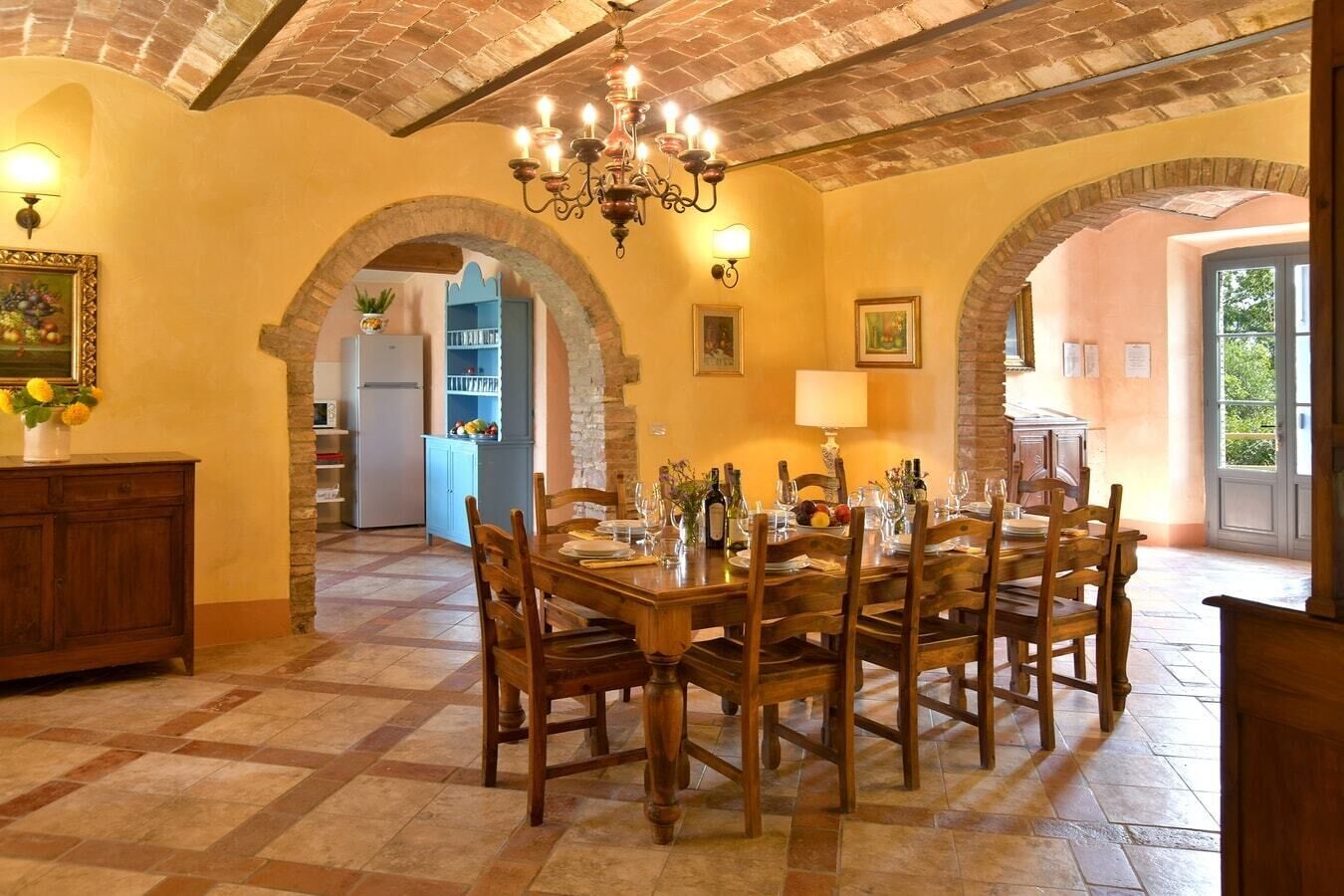 A dining area with a large wooden table set for six, surrounded by chairs. There are two archways leading to other rooms, a chandelier hanging from the ceiling, and a tiled floor.