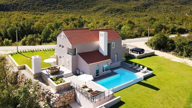 Une maison moderne à deux étages avec une piscine, entourée d’une pelouse bien entretenue et dominant un paysage montagneux.