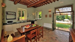 Cozinha com vigas de madeira, mesa de jantar com cadeiras, micro-ondas e vista para o jardim através de uma porta aberta.