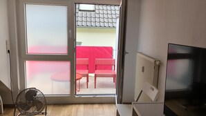 Habitación con puerta de vidrio corrediza que da a un espacio exterior con una silla roja y una mesa. Hay un radiador, un televisor y un ventilador de pie en la habitación.