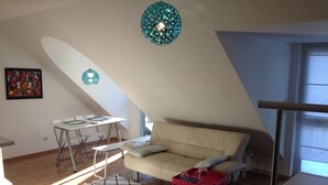 Una sala de estar moderna con techo inclinado, un sofá color beige, una pequeña mesa roja, una mesa blanca y una obra de arte colorida en la pared.