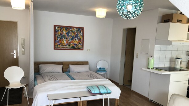 Un dormitorio moderno con una cama de madera, una mesita y una silla. Cuenta con una zona de cocina con armarios y una encimera.