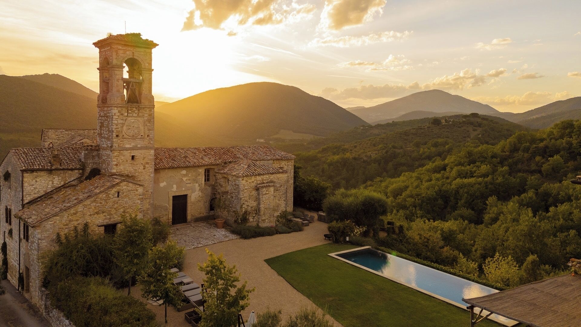 An old stone building with a bell tower, surrounded by lush greenery and mountains.
