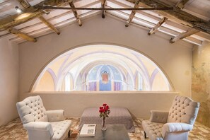 Ein Zimmer mit gewölbter Decke, einem großen Bogenfenster, zwei Polstersesseln, einem kleinen Tisch mit Buch und Blumenvase.