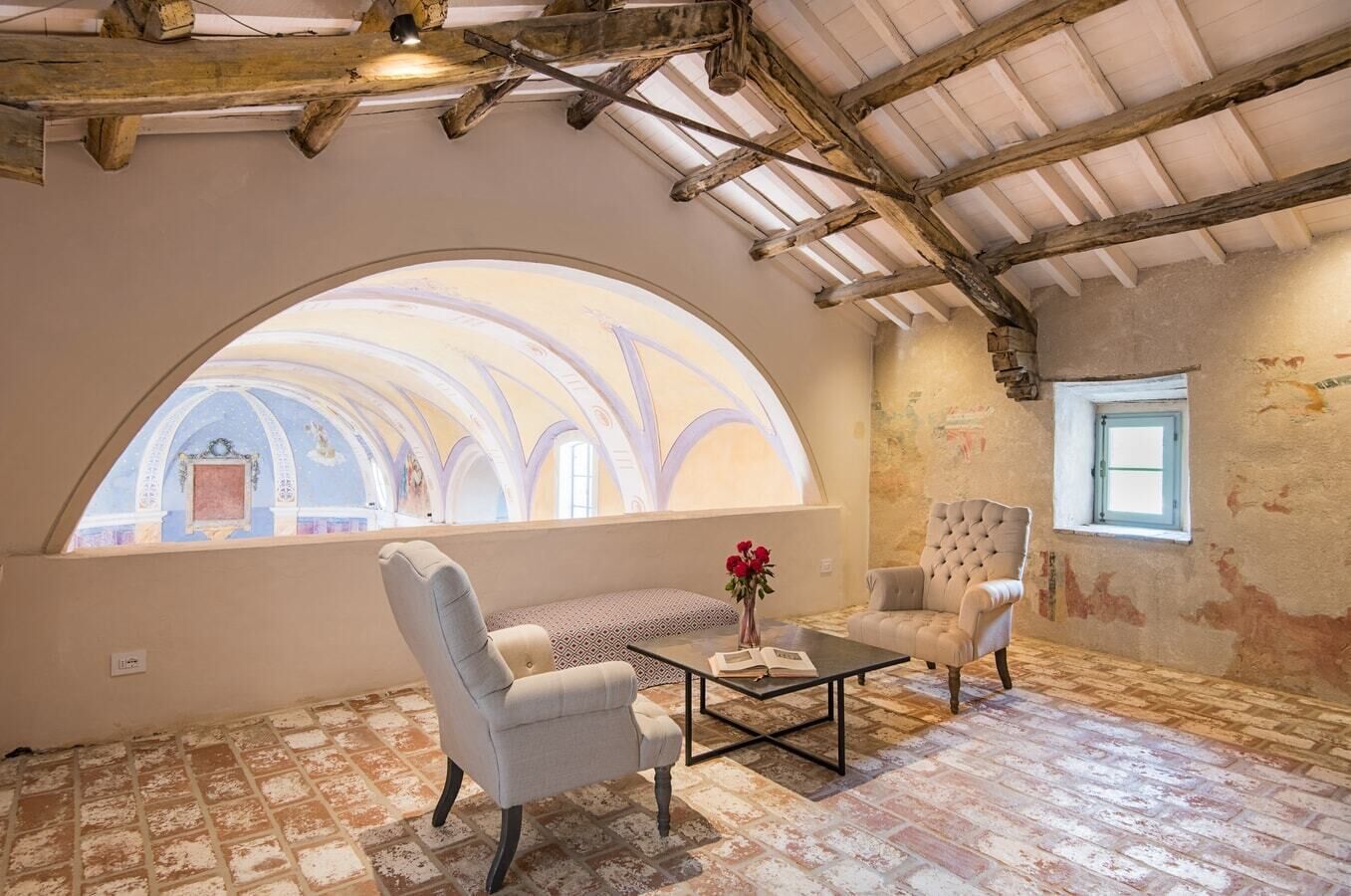 A room with a vaulted ceiling, a large arched window, two upholstered chairs, a small table, and a vase with flowers.
