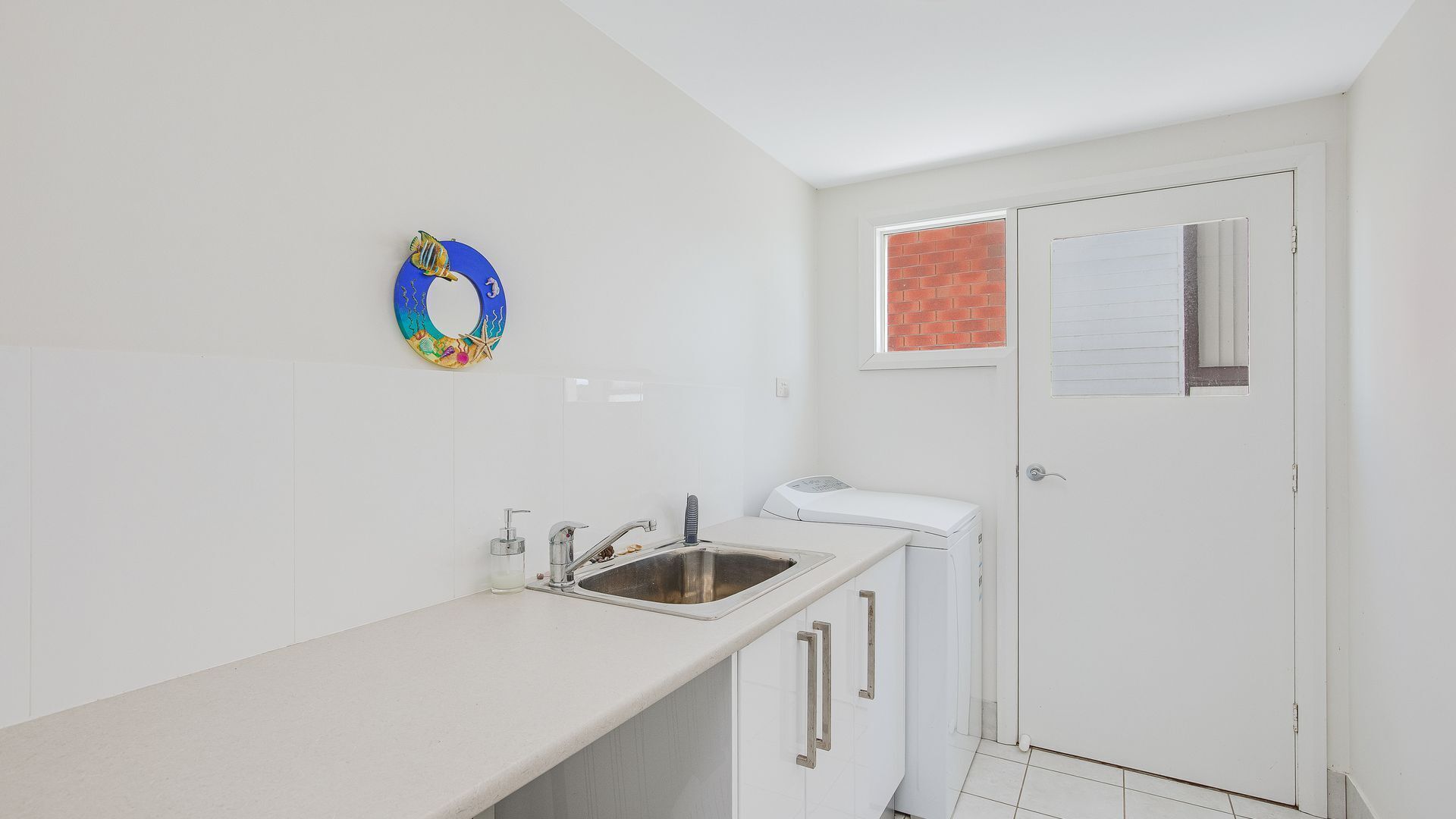 Une petite cuisine blanche, équipée d’un évier, d’un lave-linge compact et d’une porte avec une fenêtre.