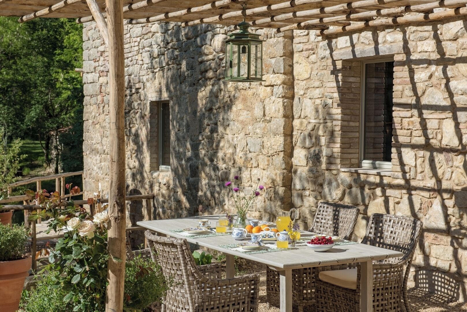 Un espace de restauration en plein air, avec un mur en pierre servant de décor, des chaises en osier et une table dressée avec une carafe de jus d’orange, un saladier de fruits et un vase de fleurs.