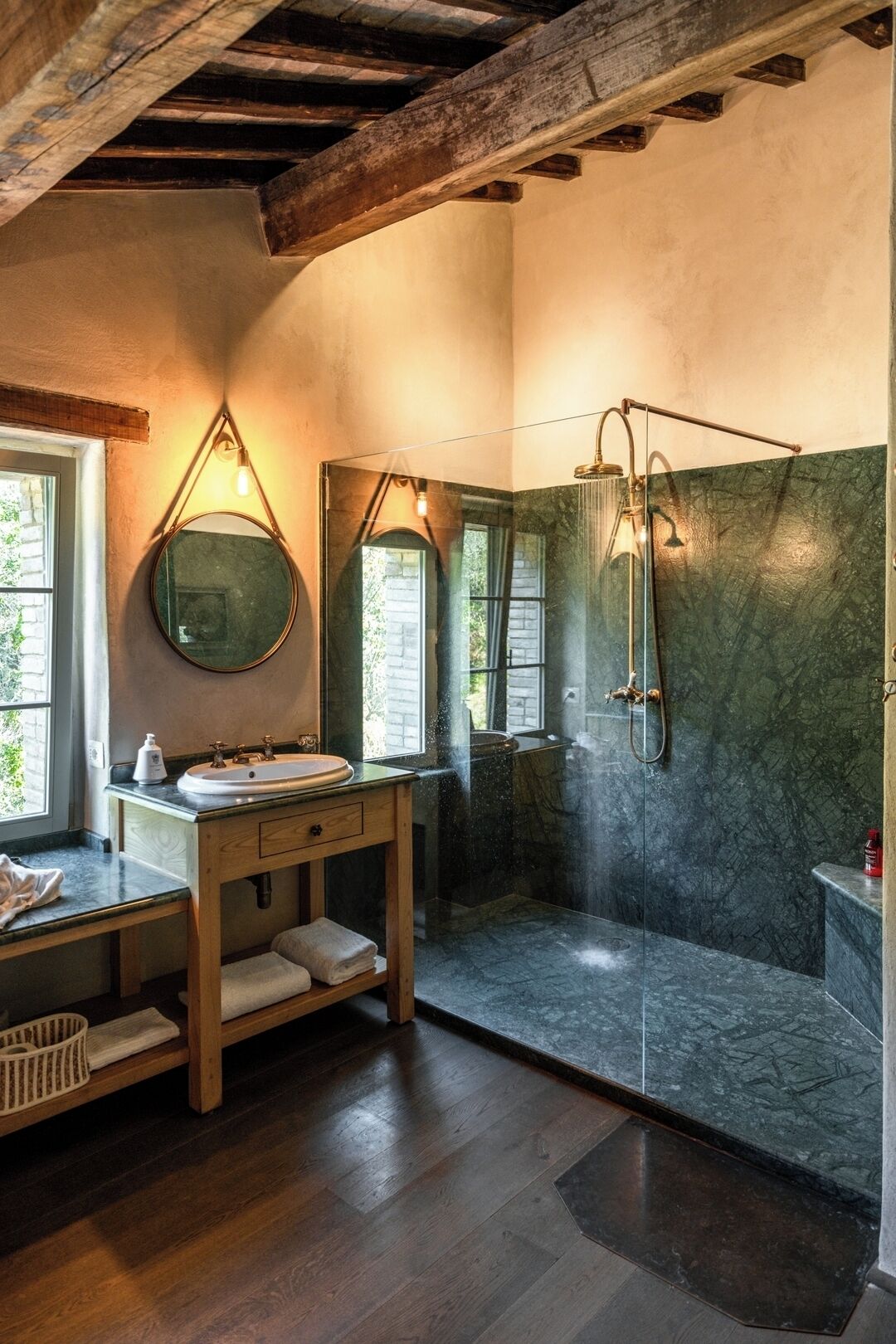 Une salle de bain avec un meuble-lavabo en bois, un miroir rond, une douche avec une porte en verre et une vue sur la verdure à l’extérieur.