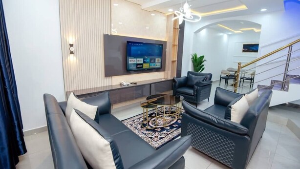 A modern living room with a flat-screen TV, leather sofas, a coffee table, and a staircase. - Iris-gold apartments (Ibadan)