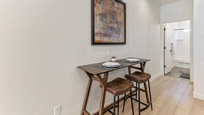 A dining area with a wooden table and two stools, a framed abstract painting on the wall, and a white door leading to a bathroom.
