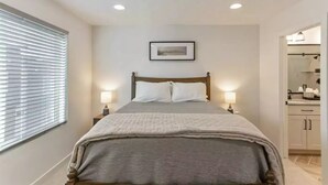 A bedroom with a bed, bedside lamps, a framed picture on the wall, and a bathroom visible through an open door.