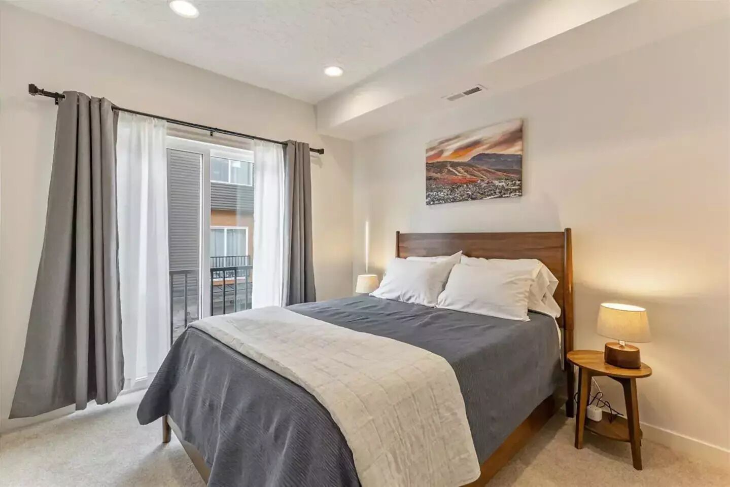 Een slaapkamer met een bed, een nachtkastje, een lamp, een raam met gordijnen en een schilderij aan de muur.