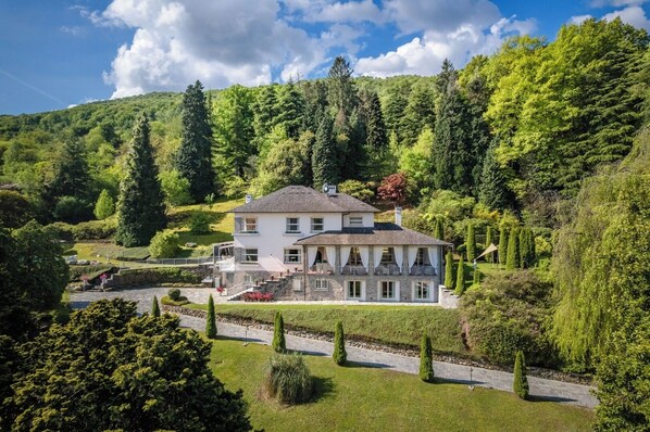Une grande maison à deux étages, dotée d’une façade en pierre, entourée d’une végétation luxuriante et d’un jardin bien entretenu.