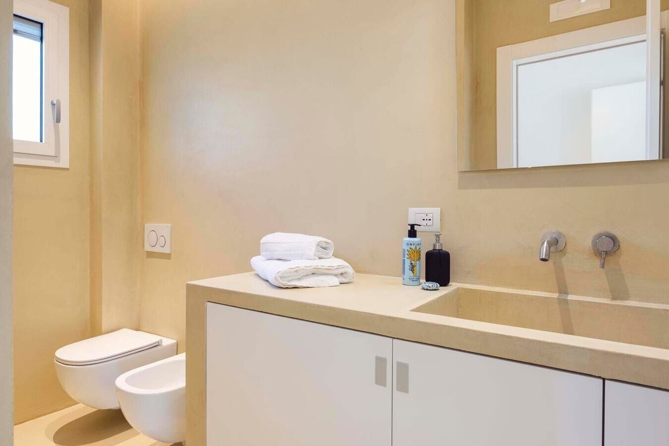Un baño moderno con inodoro, lavabo y espejo. Hay toallas y artículos de aseo en la encimera.