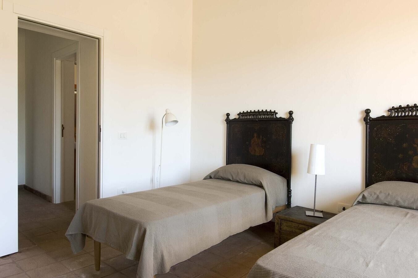 A room with two single beds, a nightstand with a lamp, a door, and a wall-mounted light fixture.