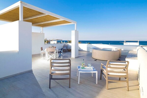 A rooftop terrace with white furniture, a wooden table, and a view of the sea.