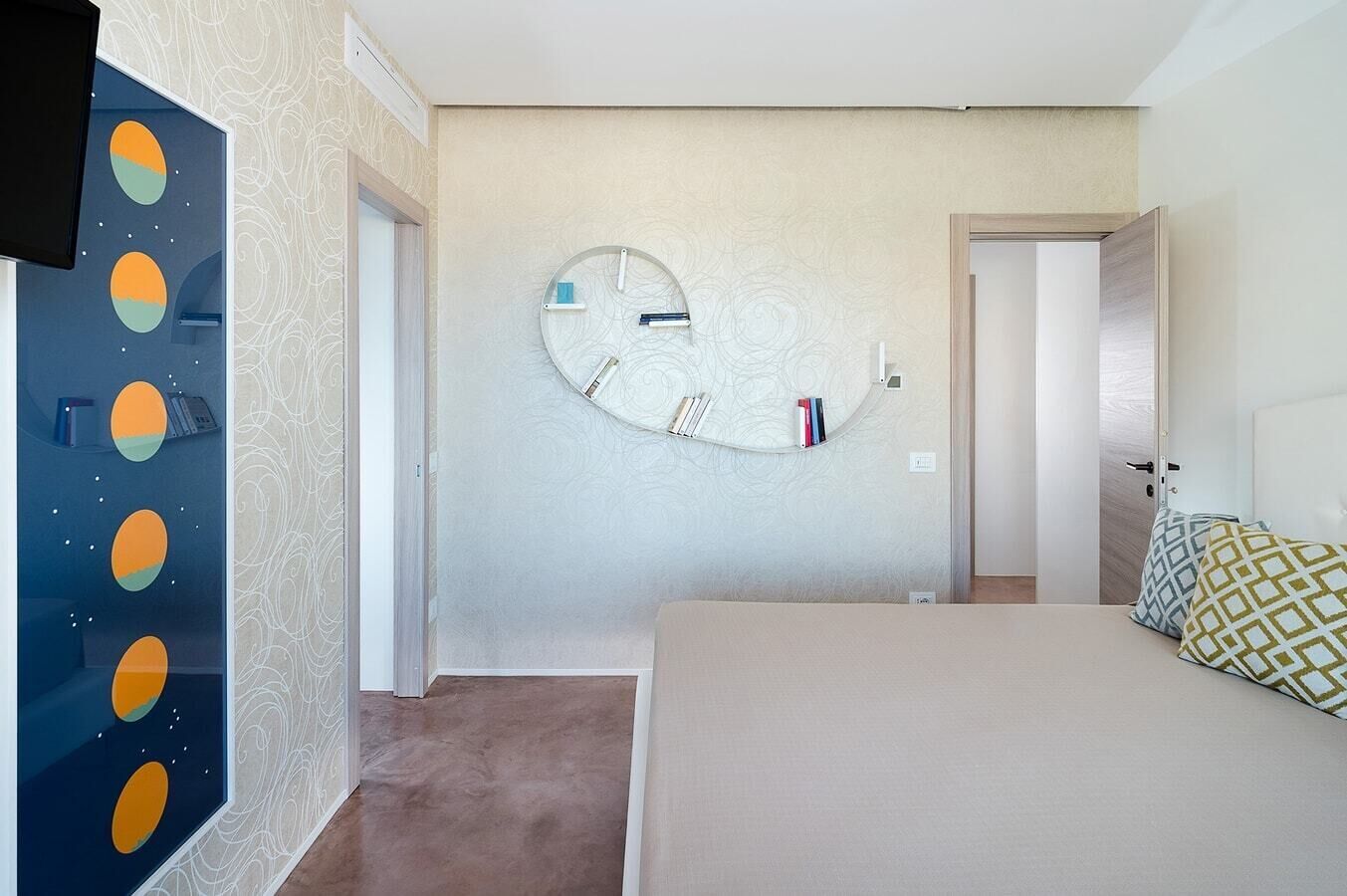 Een hotelkamer met een ronde wandklok, een bed met gestreepte kussens, een aan de muur bevestigde televisie en een deur die naar een andere kamer leidt.