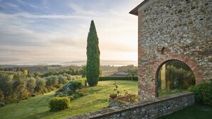A stone building with an arched doorway, surrounded by a well-maintained garden and a tall cypress tree.