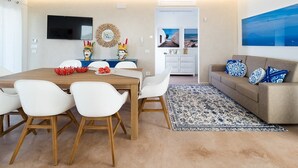 Una sala de estar moderna con una mesa de comedor de madera, sillas blancas, un sofá beige con cojines estampados en azul y blanco, un tapete con estampado azul y blanco y un televisor de pantalla plana montado en la pared.