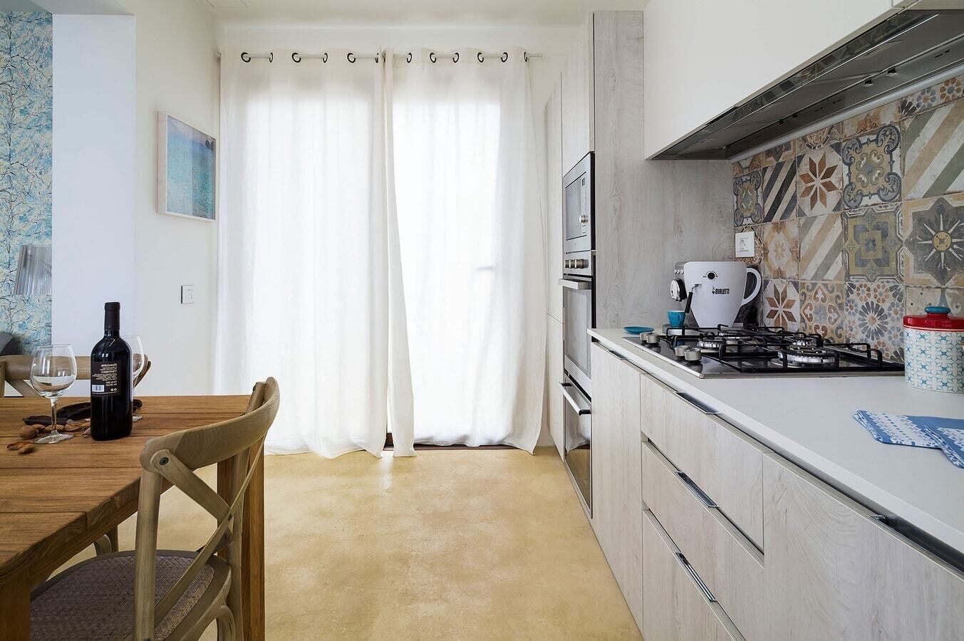 A modern kitchen with a gas stove, oven, and a coffee machine. There is a dining table with chairs and a bottle of wine.
