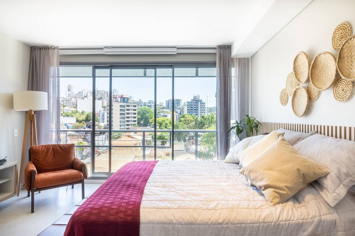 Ein modernes Schlafzimmer mit einem großen Bett, einem Sessel, einer Lampe und Blick auf die Stadt durch große Fenster.
