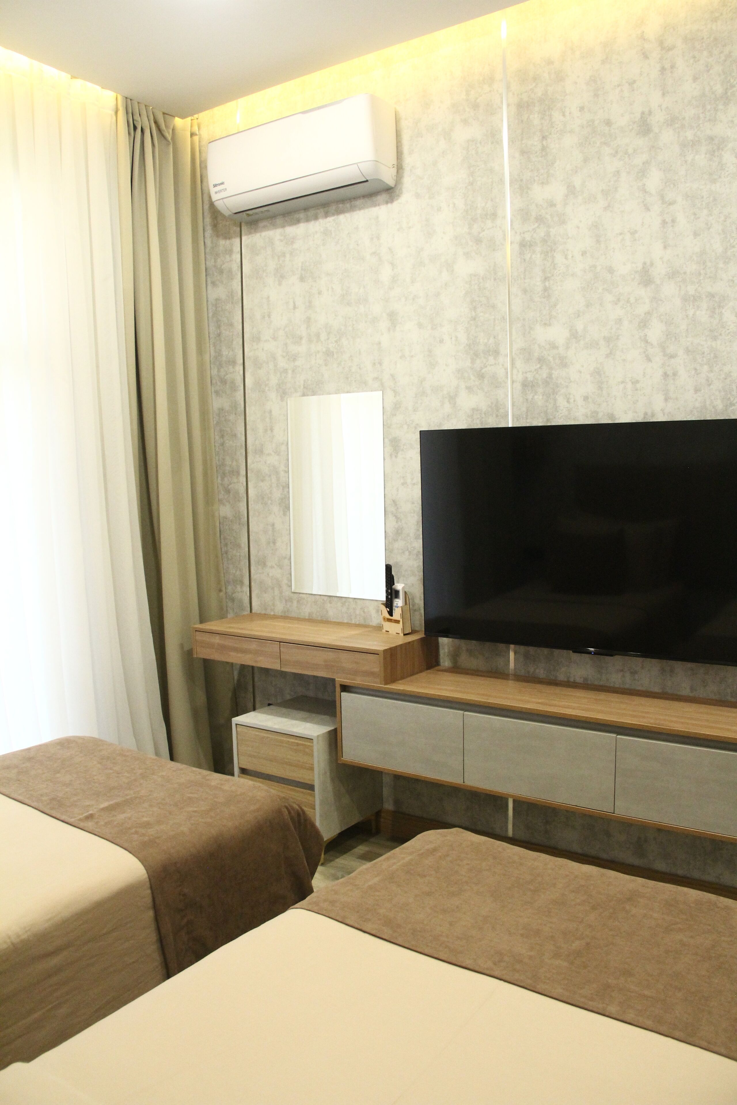 A hotel room with a flat-screen TV mounted on a wall, a wooden bedside table, a small drawer, and two beds with beige bedding.