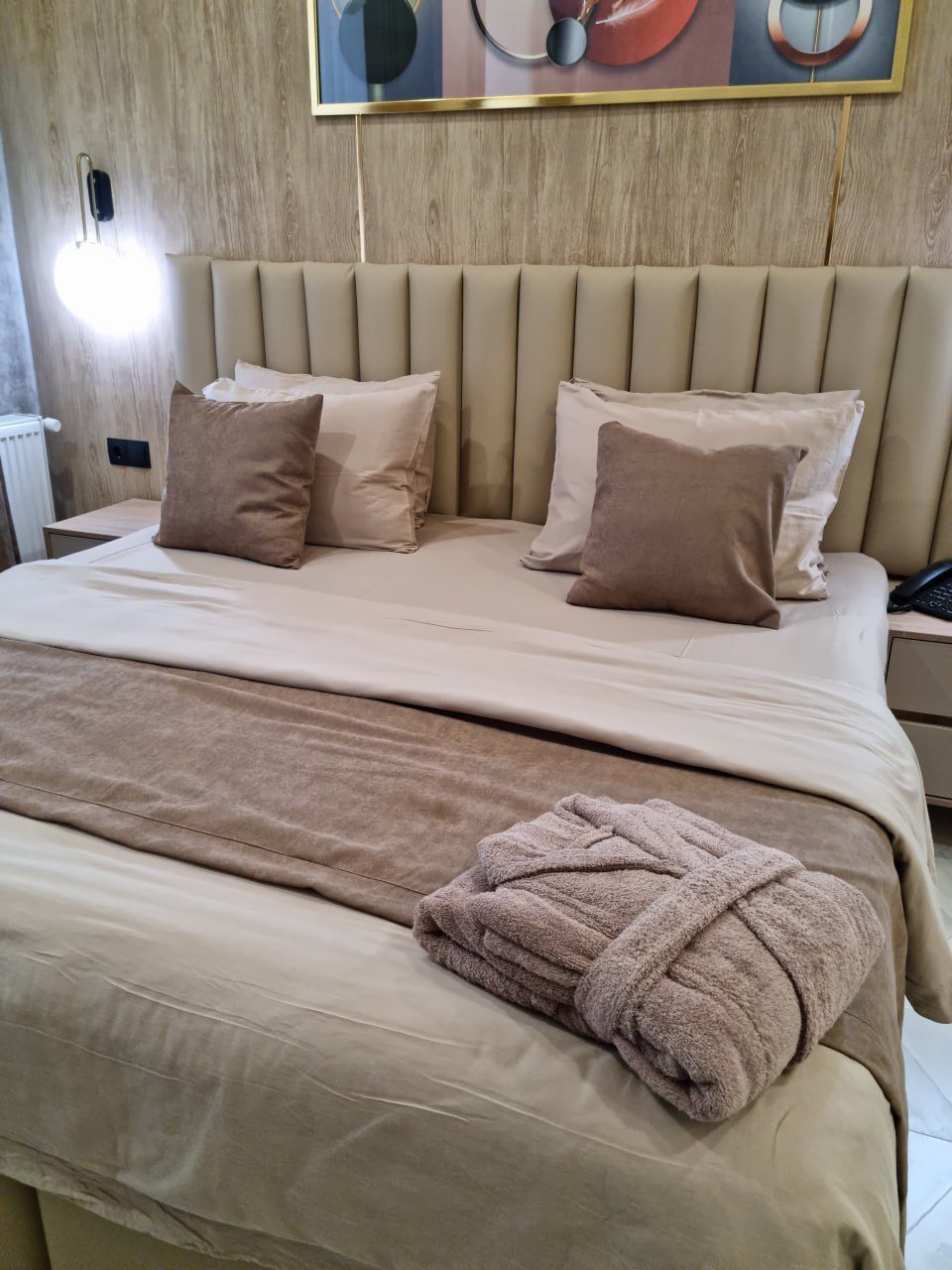 Ein ordentlich gemachtes Bett mit beiger Bettwäsche und Kissen, ein Nachttisch mit Lampe und ein an der Wand hängendes Kunstwerk.