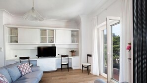 Una sala de estar moderna con un sofá gris, un televisor de pantalla plana y un comedor con armarios blancos y una silla negra.
