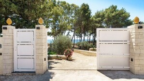 A gated entrance with a stone wall and a view of a landscaped area with trees and a clear sky. - Casa Sul Mare - Sea front house (Punta Milocca)