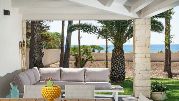 A covered outdoor seating area with a sofa and table, overlooking a beach with palm trees and the sea. - Casa Sul Mare - Sea front house (Punta Milocca)