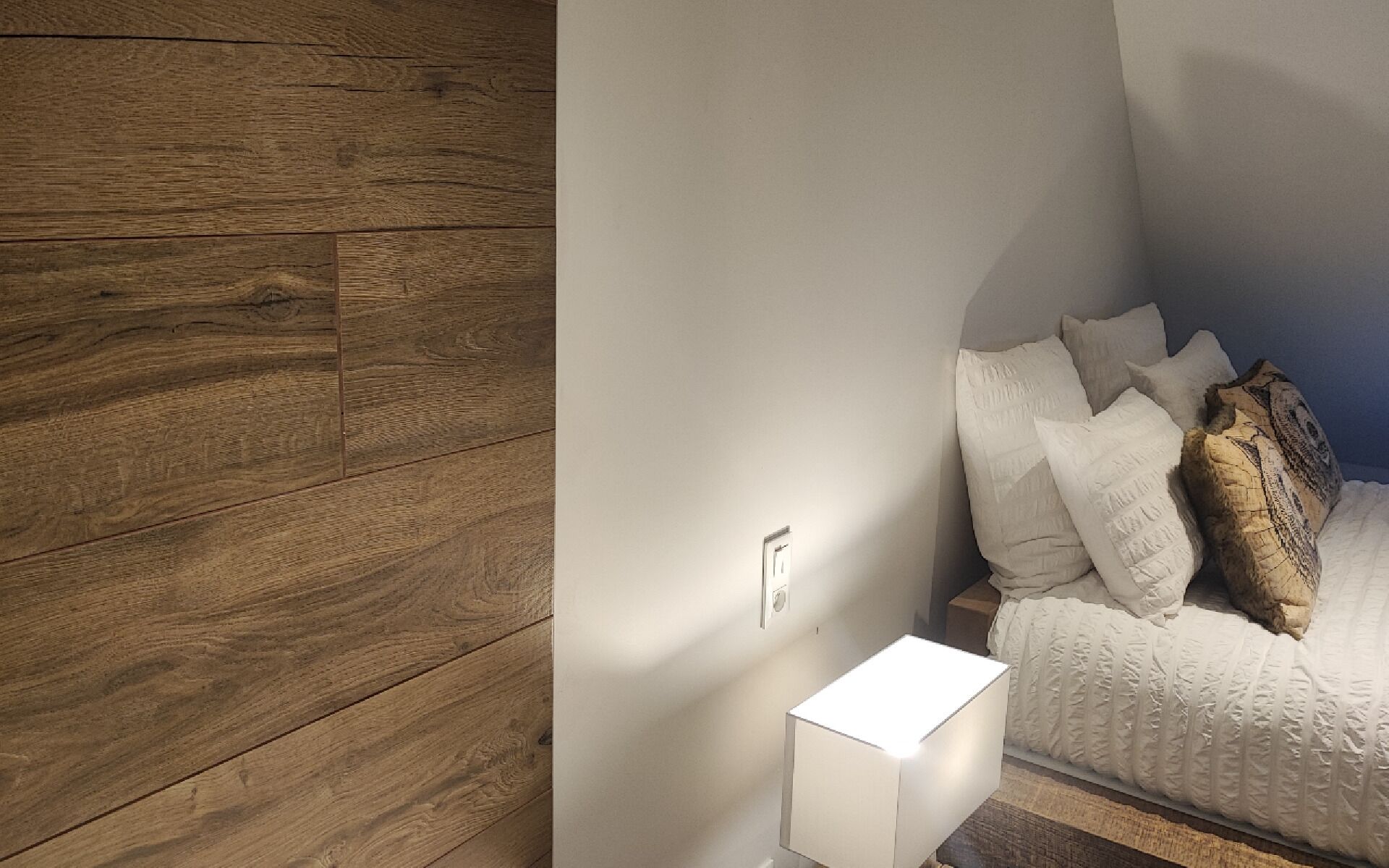 Ein Schlafzimmer mit Holzwand, Nachttisch mit Lampe und einem Bett mit vielen Kissen.