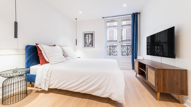 A modern bedroom with a bed, a TV mounted on the wall, a small table, and a window with curtains.